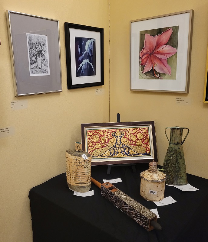 Three framed floral pictures hanging on a yellow wall. A small table with a black tablecloth sits below these paintings. The table holds 4 pottery pieces and a framed picture on an easle.